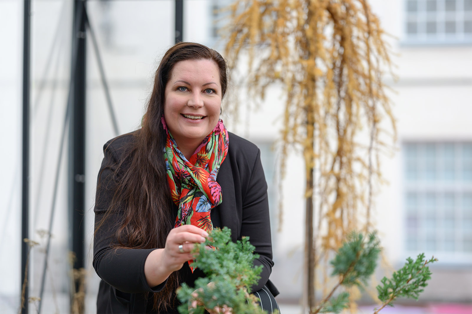 Nainen hymyilee suoraan kameralle ja koskettaa edessä olevaa kukkaa.